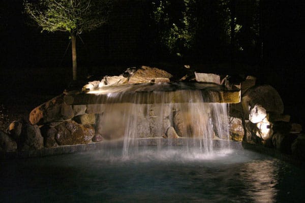 A Stone waterfall grotto in a backyard pool illuminated by lighting